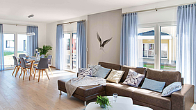 Wohn-Esszimmer brauner Holzfußboden weiß und graue Wände bodentiefe Fenster blaue Gardinen braune Couch mit blauen Kissen Esszimmertisch mit 6 grauen Polsterstühlen